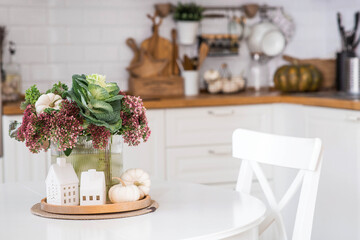 Still-life. A bouquet of decorative cabbage in a vase, pumpkins and white houses on the table in the interior of a home kitchen. A cozy autumn concept.