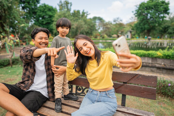 young mom takes cell phone selfie with father and little boy while sitting on park bench