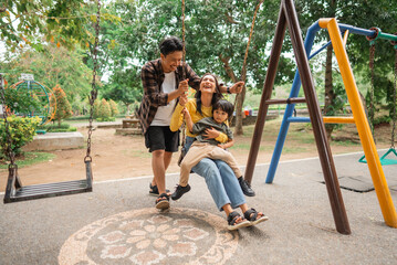 young woman holding little boy on her lap playing swing with young man in park