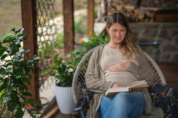 Portrait Caucasian pregnant mature relaxed woman sitting in chair on cottage terrace and reading book, gently touching belly