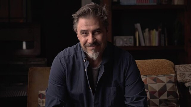 Portrait of older man at home in dark room sitting on couch. Happy smile, grey hair, bearded, . mature age, middle age, mid adult man in 50s.