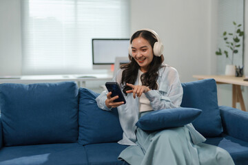 Young woman is sitting on a blue sofa at home, wearing headphones and smiling while using her smartphone