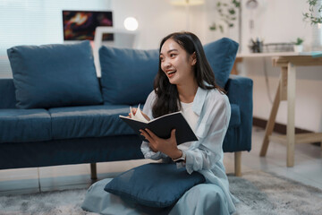 Young asian woman is sitting on the floor in her living room, taking notes in a notebook and smiling