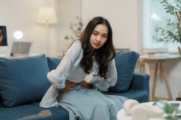 Young woman sitting on her sofa and holding her stomach in pain, suffering from menstrual cramps or...