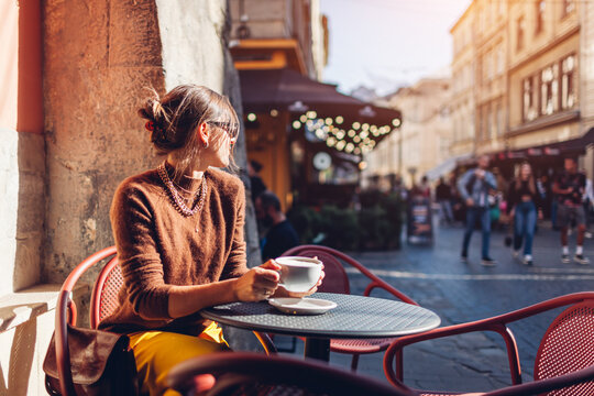 Portrait of stylish woman sitting in cafe outdoor drinking coffee. Woman enjoying architecture of Lviv city on street