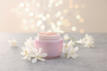Cream in jar and jasmine flowers on grey table against beige background with blurred lights, closeup