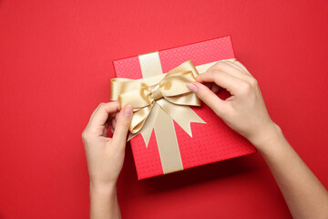 Woman decorating gift box with bow on red background, top view