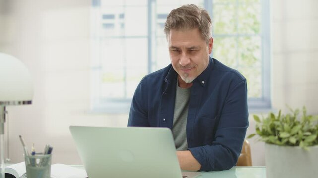 Happy mid adult man working with laptop computer at home, sitting at desk with confident smile. Businessman online form home office, Portrait.