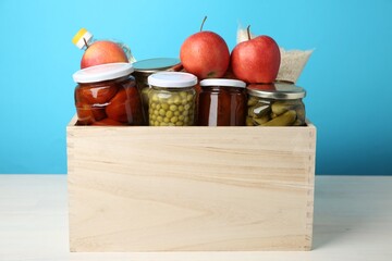 Different food products for donation in wooden crate on table against light blue background