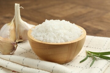 Sea salt in bowl, rosemary and garlic on brown table, closeup