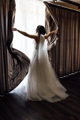 Fototapeta premium The girl stands with her back near the window and opens the curtains with her hands