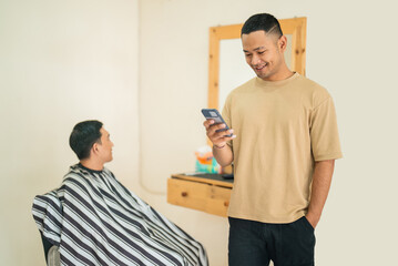 Obraz premium A man is standing closely next to another man who is seated in a barber chair, intently looking at his cell phone and checking messages