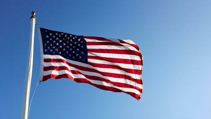 Obraz premium The stars and stripes of the American flag flutter gracefully in the wind against a bright blue sky, a perfect symbol of unity and patriotism