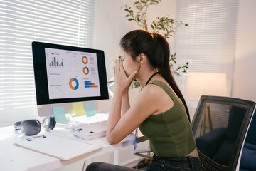 Young businesswoman is suffering from stress and headache while working on a laptop at the office. She is sitting at her desk with graphs and charts on the computer screen