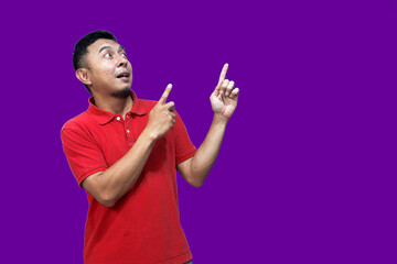 An excited Asian man wearing a red shirt points to the empty space above him, isolated on a purple background.