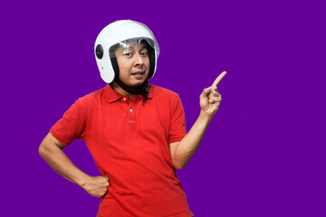 Naklejka premium Young Asian driver man wearing helmet looking up while raising one hand and pointing upwards with happy expression 