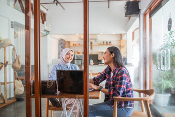 two laughing Indonesian women sit using a laptop computer together in a coffee shop