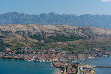 Aerial view of a coastal town with mountain backdrop, red-roofed buildings, and a vibrant seaside beach. Summer day in a Mediterranean coastal town surrounded by rugged hills and clear blue waters.