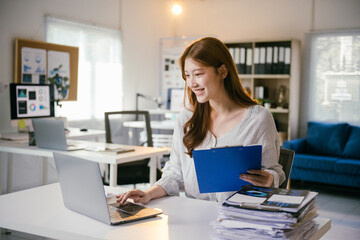 Obraz premium Smiling asian businesswoman works on her laptop in the office, analyzing marketing data and handling paperwork with a focus on corporate success