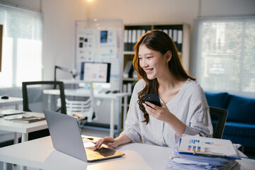 Smiling businesswoman is working on a laptop and using a smartphone at her desk in a modern office. She is surrounded by paperwork and digital devices