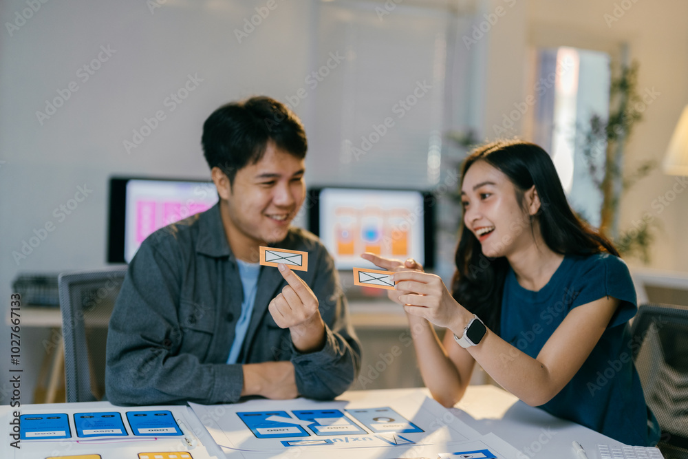 Aufkleber Two asian ui/ux designers smiling and discussing the design of a mobile application interface during a meeting in the office