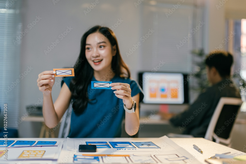 Aufkleber young asian woman working as ui/ux designer is smiling while holding two colorful paper prototypes d