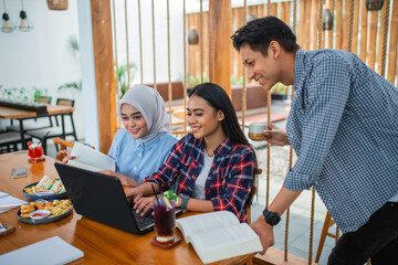 Fototapeta premium Group of Asian students studying together on a laptop while enjoying a drink at a coffee shop