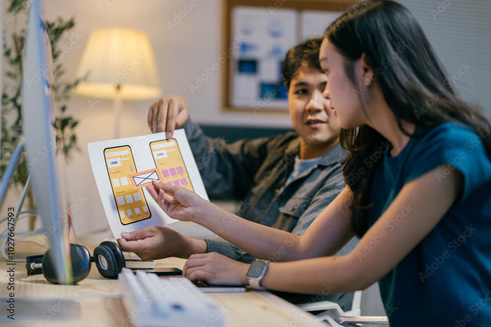 Poster Two asian ui/ux designers collaborating in the office, analyzing and discussing a mobile app interface design, showcasing teamwork and dedication for their startup