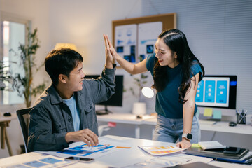 Two coworkers are giving each other a high five over a table with paperwork and digital devices, celebrating a successful business project