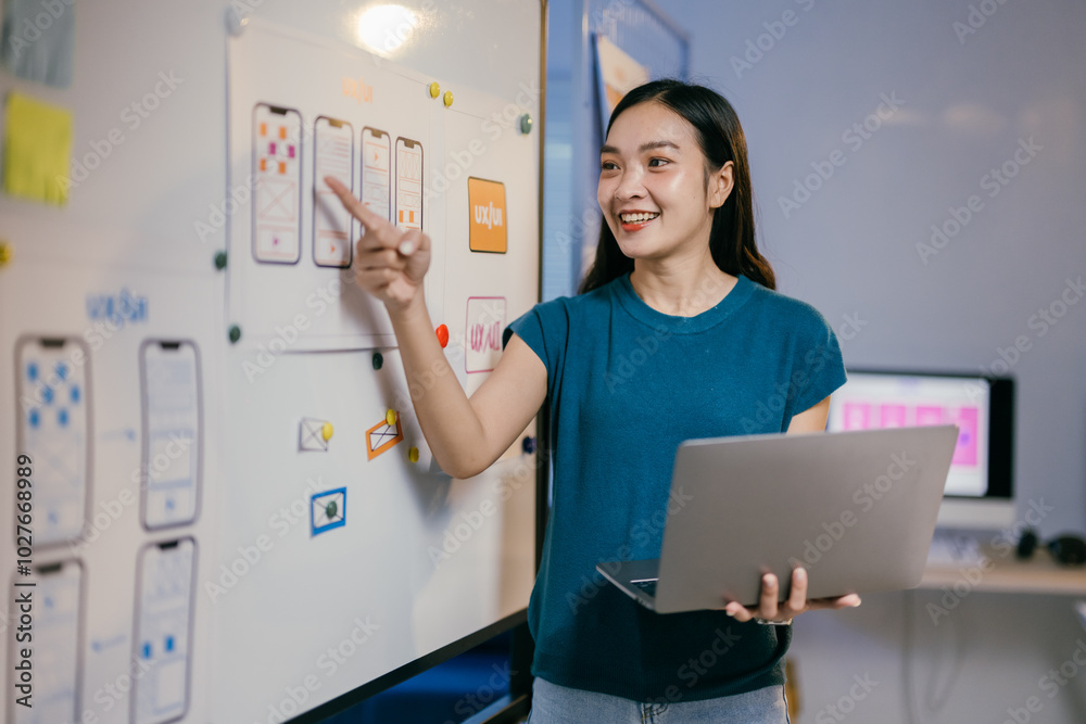 Poster Ui ux designer is standing and pointing at a whiteboard with mobile application interface wireframes on it while holding a laptop