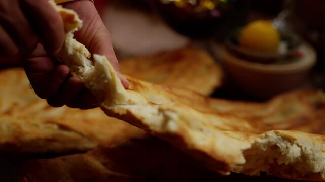 Close-up of a baker breaking a freshly baked flatbread. The baker is wearing a yellow shirt and has his hands covered in flour