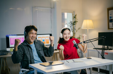 Obraz premium Two asian content creators are recording a podcast together, wearing headphones and cheering with their arms raised while sitting at a table with a microphone