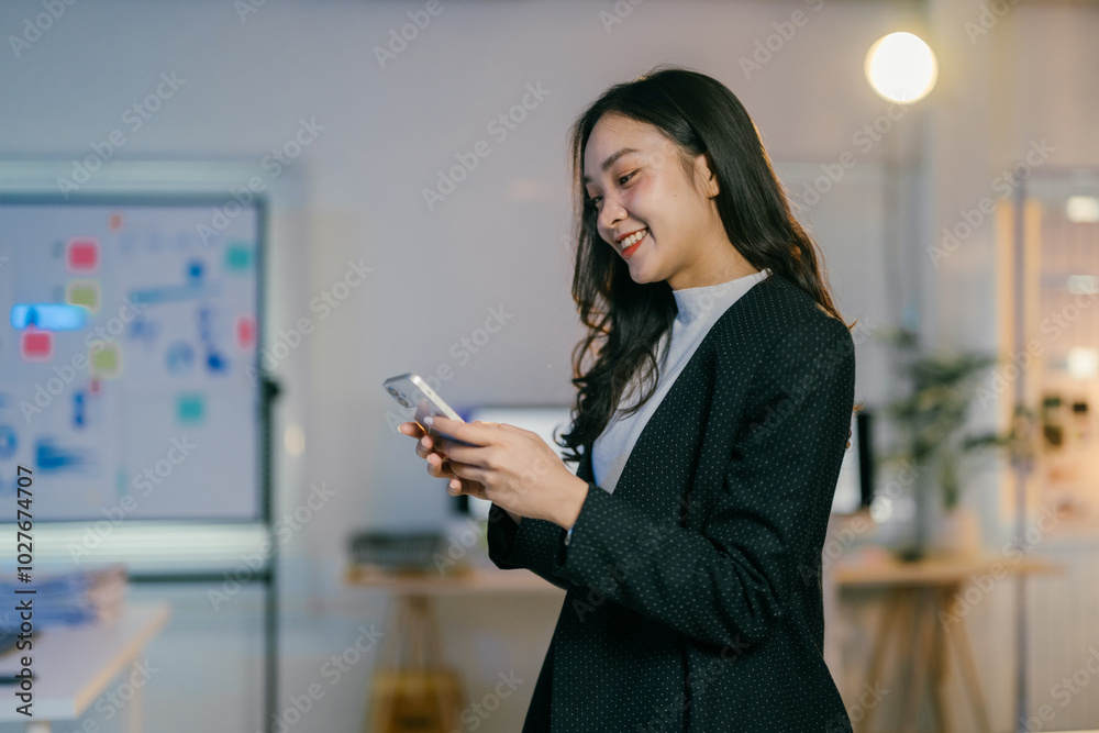 Poster Young manager is smiling while using her smartphone in a modern office with a whiteboard and sticky notes in the background