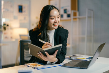 Focused businesswoman smiles and writes in her notebook during a late-night video call,...