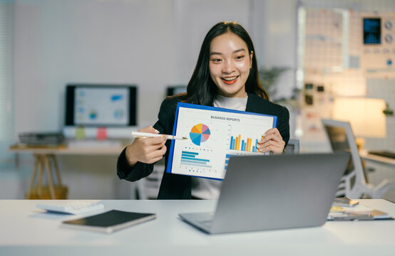 Young businesswoman is holding charts and pointing at data while having a video call on her laptop, explaining statistics, and discussing business reports with colleagues