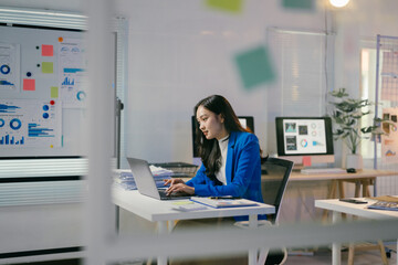 Young asian businesswoman is working on her laptop in a modern office, surrounded by charts and...