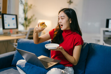 Young asian woman is sitting on a blue sofa, eating a burger and watching a movie on her laptop. She is holding a plate in one hand and a burger in the other