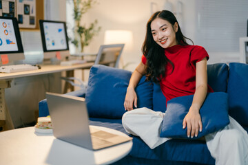 Obraz premium Young woman with long dark hair is sitting cross-legged on a blue sofa and watching something on her laptop with a big smile
