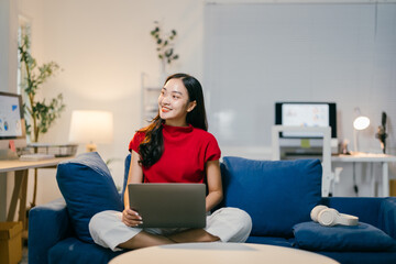 Young asian woman happily works on her laptop in her cozy home office, surrounded by modern design and natural light. She is fully immersed in her work, dressed casually and smiling