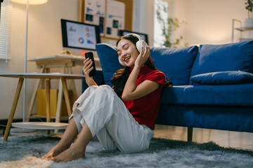 Young woman is enjoying her free time at home, relaxing on the floor while listening to music on her headphones and browsing on her phone