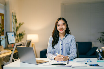 Dedicated businesswoman works late in the office, surrounded by paperwork and a laptop, exuding confidence and cheerfulness as she focuses on her tasks