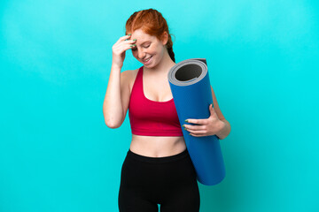 Young sport reddish woman going to yoga classes while holding a mat isolated on blue background...