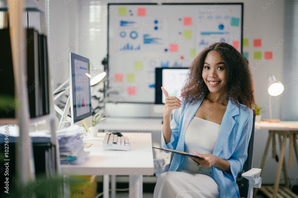 Poster Office worker is holding a tablet and gesturing thumbs up while sitting at her desk with graphs and charts on the background