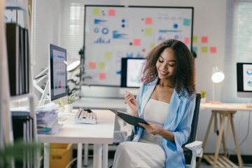 African american businesswoman is sitting at her desk reviewing documents with a thoughtful expression. With a bright smile, she appears to be content with her work