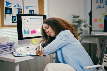 Young businesswoman is falling asleep at her desk while working on a project. She is surrounded by paperwork and has a tired expression