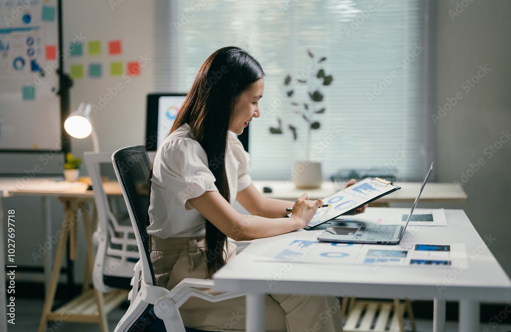 Poster asian businesswoman is analyzing financial graphs on a clipboard while sitting at her desk in the of