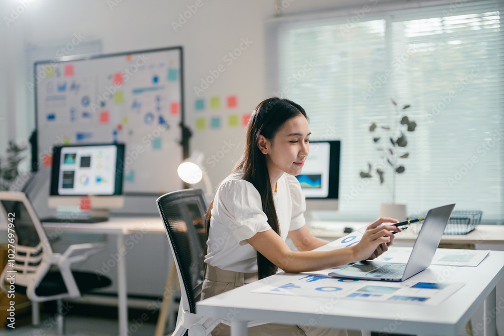 Aufkleber Businesswoman working with laptop computer and analyzing marketing information on a document with charts and graphs while sitting at her desk in the office
