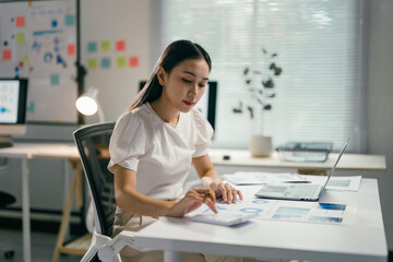 Focused young asian businesswoman analyzing financial data with a calculator in a modern office,...