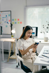 Young asian businesswoman is sitting at her desk in a modern office, smiling and using her smartphone. She is wearing casual clothing and appears to be enjoying her work