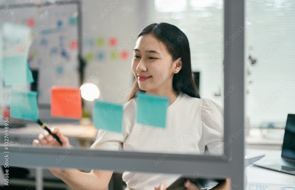 Poster young asian businesswoman is using a marker to write on sticky notes while brainstorming new project